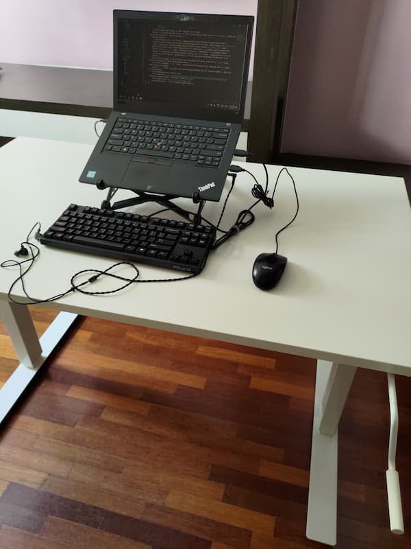 my home desk on a wooden floor with ikea standing desk, a crank on forefront, thinkpad on a roost stand, in-ear earphone and mouse and keyboard plugged to the laptop's ports